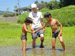 画像：「教育ファーム」でどろんこ相撲をする子どもたちと「みつめよう」のマークのＴシャツを着た浅川氏